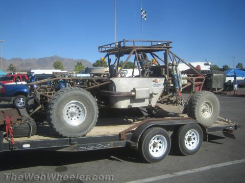 1990 Ford Bronco - bare-metal tube-frame off-road race buggy with long-travel suspension sits loaded on a tandem-axle flatbed trailer in a lot.