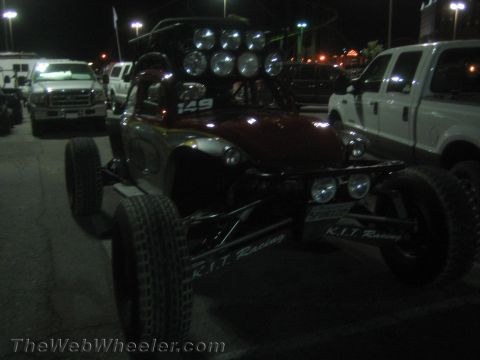 1990 Ford Bronco - dark open tube-frame off-road race buggy with a roof light bar marked K.I.T. Racing sits between Ford pickups in a lot at night.