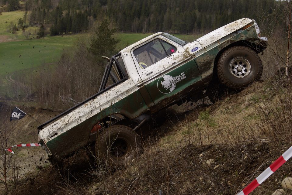 White and green Ford pickup truck climbing a muddy hill with off-road tires and a roll cage