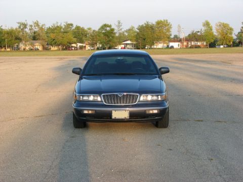 1985 Mercury Grand Marquis - A dark sedan is parked in an open lot during the day, with a suburban background.