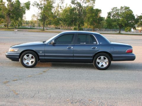 1985 Mercury Grand Marquis - A blue sedan is parked on an asphalt lot with trees and a fence in the background.