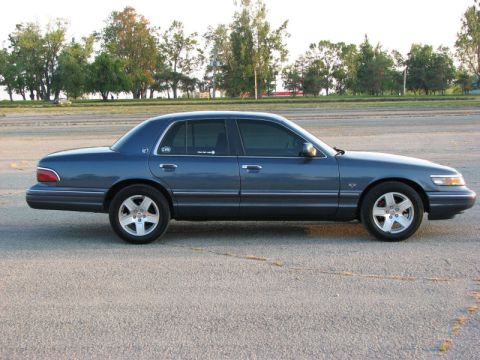 1985 Mercury Grand Marquis - This blue sedan, likely a 1998 model, is shown from the side against a backdrop of trees and a road.
