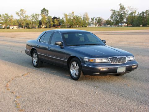 1985 Mercury Grand Marquis - A dark blue sedan is parked on a gravel surface with trees and houses in the background.