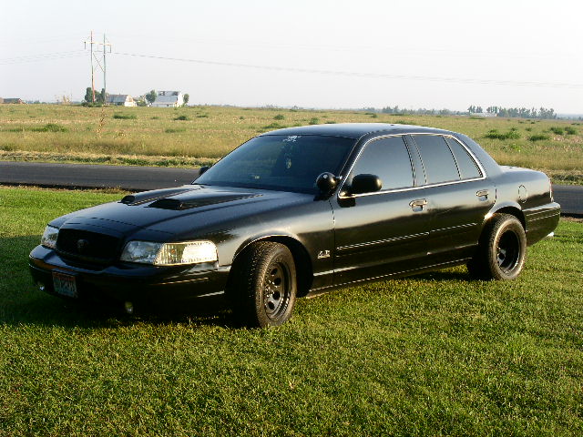 Front three-quarter view of a black Crown Victoria sedan with hood scoop and black steel wheels on grass