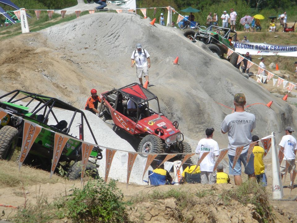 Red tube-chassis rock crawler climbing a dirt hill at a competition with spectators.