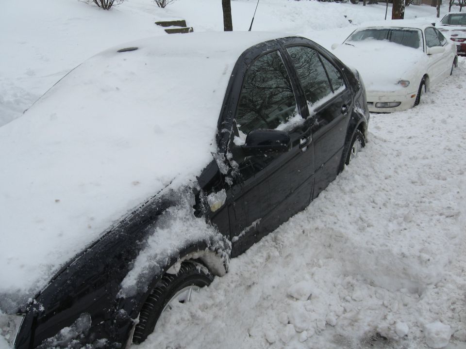 Black Jetta sedan buried in deep snow at curb, with snow-covered white car parked behind it