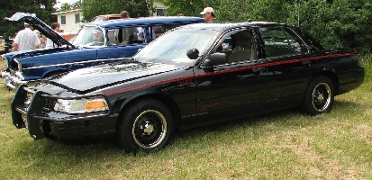 2000 Ford Crown Victoria - Black 2000 Ford Crown Victoria with a push bumper is parked on grass next to a blue vintage car.