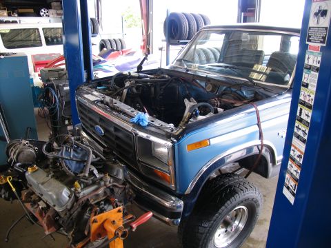 1984 Ford Bronco - The engine bay of a blue truck is shown with the engine removed and placed on an engine stand for repair.