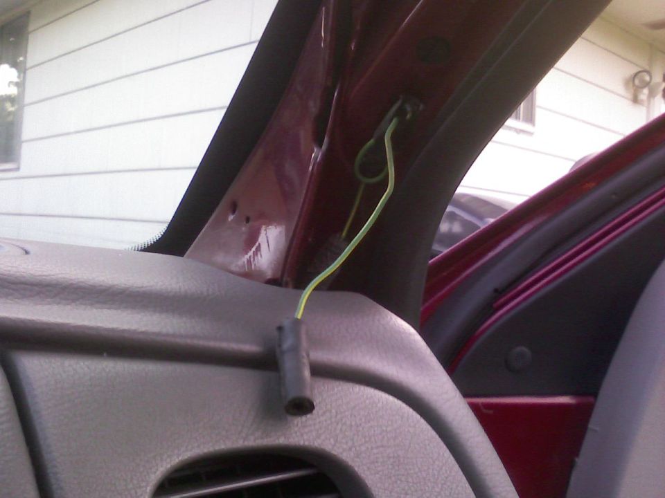 Interior view of a car's A-pillar with exposed green wiring and a black connector. The dashboard and door frame are visible
