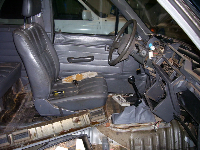 Interior of a vehicle with seats and dashboard removed, showing exposed metal and wiring. Another white vehicle is visible in the background