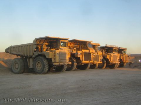 1968 Ford Mustang - Several large yellow mining dump trucks are parked in a line on a dirt lot under a clear sky.