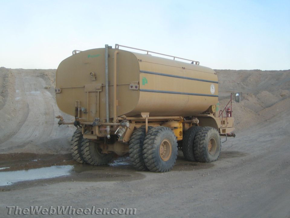 Rear view of a tan water truck with large tires parked on a dirt road