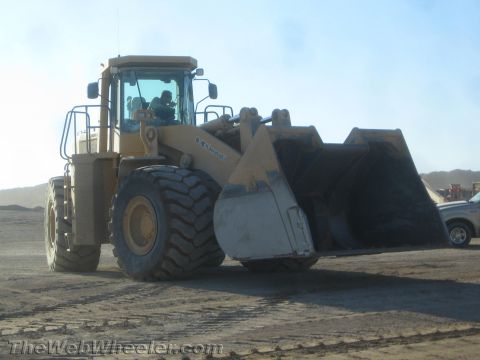 1968 Ford Mustang - A large yellow wheel loader is parked on a dirt surface under a clear sky. Another vehicle is partially visible to the right.