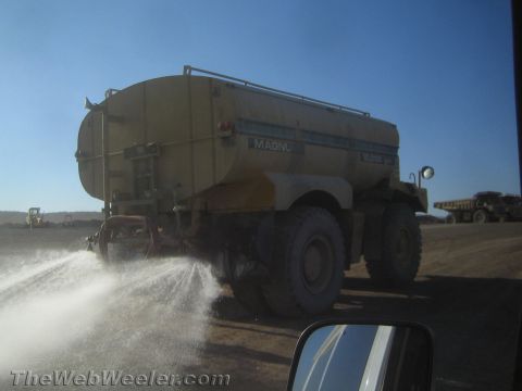 1968 Ford Mustang - A large yellow water truck sprays water on a dusty surface, with another heavy vehicle visible in the distance under a clear blue sky.