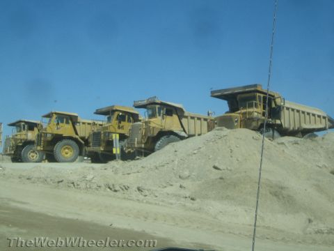 1968 Ford Mustang - Several large yellow dump trucks are parked in a line on a dirt lot under a clear blue sky.