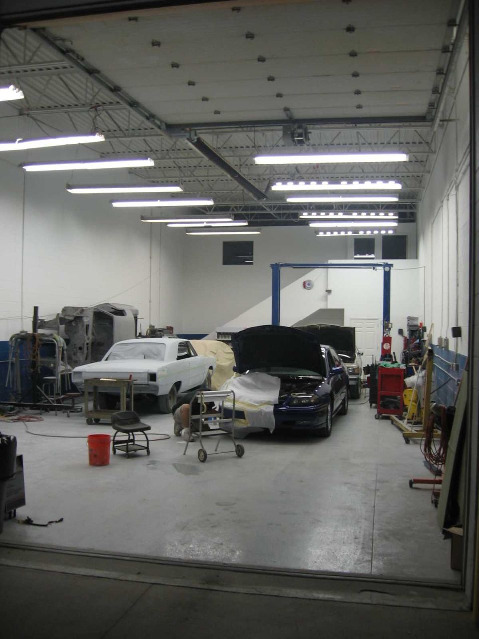 Two cars are in an auto repair shop, one is white and covered, the other is dark blue with its hood open