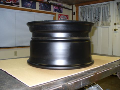 1978 Ford Bronco - A black metal wheel rim sits on a workbench, resting on a piece of cardboard. The background shows a garage-like setting.
