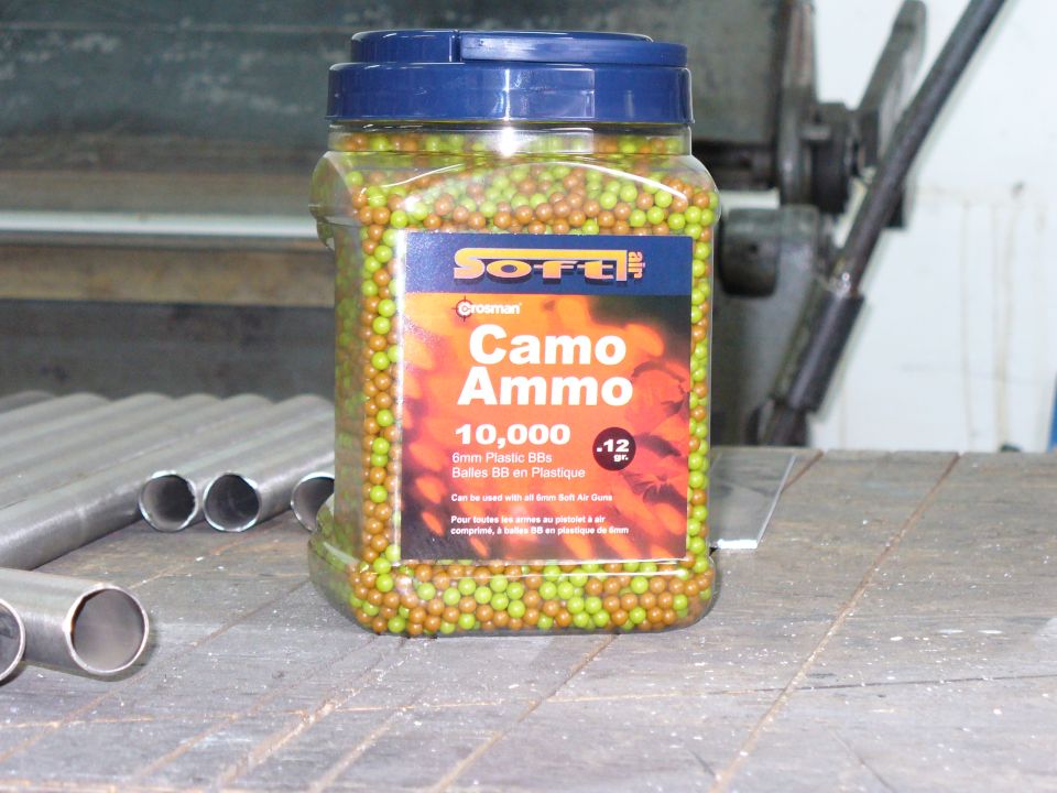 Jar of 10,000 6mm plastic BBs labeled Camo Ammo, on a wooden surface with metal pipes