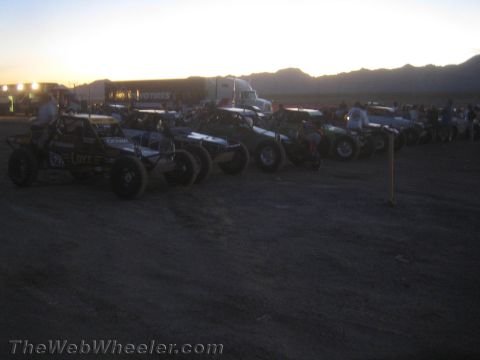 1990 Ford Bronco - line of off-road race buggies and trucks staged in a dirt lot at dusk, silhouetted against a hazy mountain skyline and big rigs.