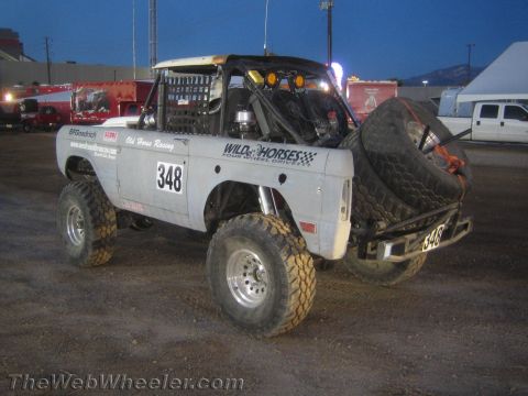1990 Ford Bronco - White early Ford Bronco off-road race truck #348 with a rear-mounted spare tire and Wild Horses graphics parked near buildings at dusk.