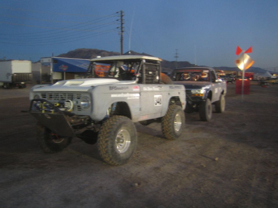 White Ford Bronco race truck #348 and blue Ford truck driving in dirt at dusk