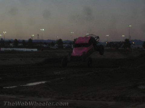 1990 Ford Bronco - pink-bodied off-road race buggy launches off a dirt jump against a dusky sky with stadium lights in the background.