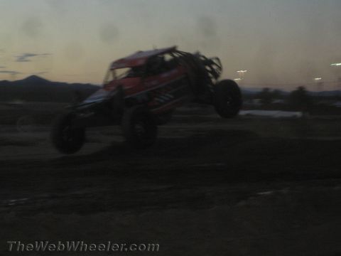 1990 Ford Bronco - red and black tube-frame off-road race buggy catches air over a dirt course jump at dusk with mountains in the distance.