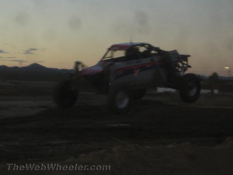1990 Ford Bronco - white and red off-road race buggy leaps a dirt jump at dusk, all four wheels off the ground against a hazy mountain backdrop.
