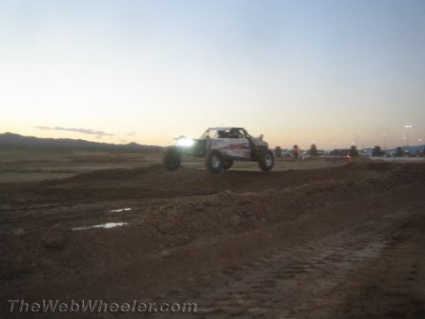 1990 Ford Bronco - off-road race truck with headlights blazing catches air over a dirt course berm at dusk with mountains on the horizon.