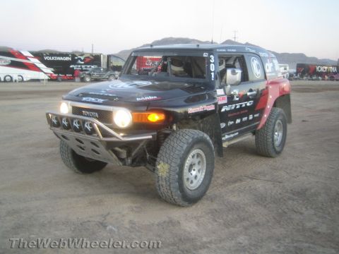 1990 Ford Bronco - Black Toyota FJ Cruiser off-road race truck #301 with Nitto graphics and headlights on, staged in a dirt pit near transporters.
