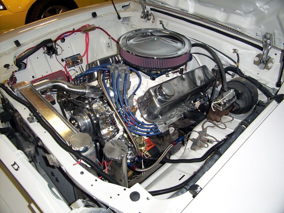 Engine bay showing a V8 engine with chrome valve covers, air cleaner, and custom wiring. A yellow car is partially visible in the background
