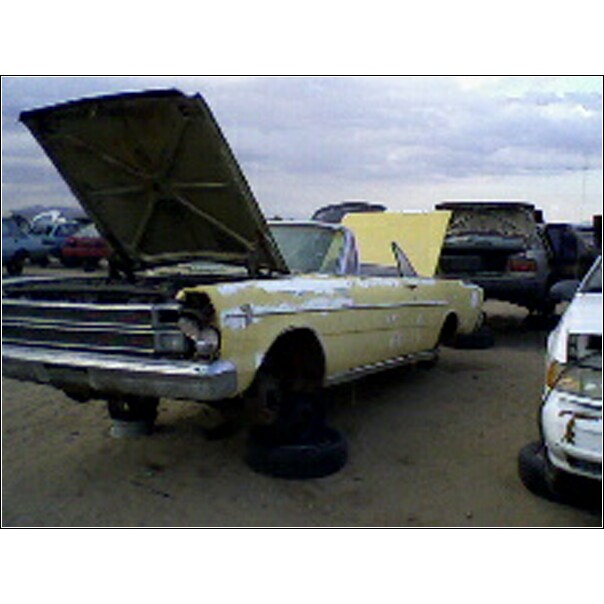 Yellow convertible with hood up, sitting on tires in a junkyard. Other cars visible in background