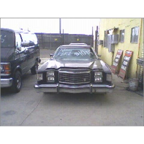 1973 Ford Ranchero - A dark sedan with a prominent grille and square headlights is parked outside a building with two other vehicles nearby.
