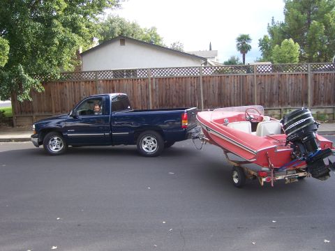 1991 Ford Bronco - A blue pickup truck is seen from the side, towing a red boat on a trailer down a residential street.
