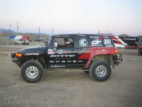 1990 Ford Bronco - Side profile of a black and red Toyota FJ Cruiser off-road race truck #301 with Nitto and KC graphics in a dusty staging lot.
