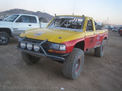1990 Ford Bronco - Yellow and red off-road race pickup #741 with a tube front bumper and round lights parked in a dusty staging lot beside a Dodge truck.