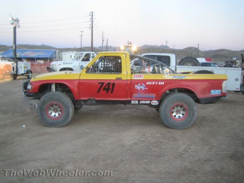 1990 Ford Bronco - Side profile of a red and yellow off-road race pickup #741 with a caged cab and bypass shocks parked at dusk among support trucks.