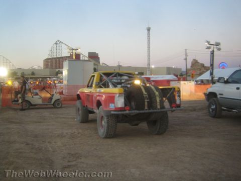 1990 Ford Bronco - Rear of a yellow and red off-road race truck carrying dual spare tires, parked in a dirt lot near a fairground with a roller coaster behind.