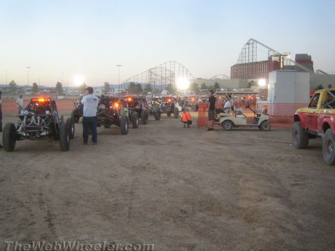 1990 Ford Bronco - Off-road race vehicles lined up on a dirt lot at dusk, with a roller coaster and stadium lights in the background.