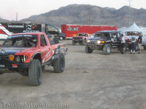 1990 Ford Bronco - Several modified off-road race trucks gathered on a dirt staging area with rugged desert mountains in the background.