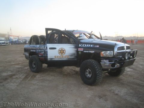 1990 Ford Bronco - black Dodge Ram off-road race truck wearing Sheriff-themed livery, a roll cage, and large tires parked on dirt.