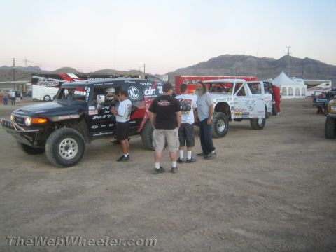 1990 Ford Bronco - group of people gather beside off-road race trucks parked on dirt with desert hills and tents in the background.