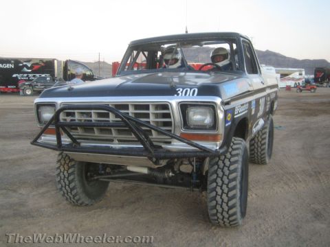 1990 Ford Bronco - blue Ford number 300 off-road race truck with a tubular front bumper and two helmeted occupants on a dirt lot.