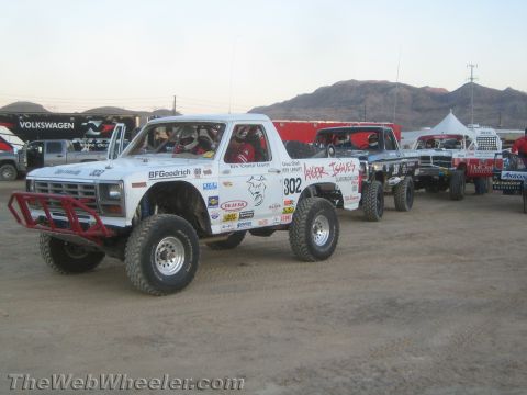 1990 Ford Bronco - white Ford number 302 off-road race truck with BFGoodrich graphics staged with other race trucks in a desert pit.