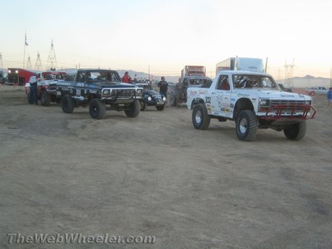 1990 Ford Bronco - row of off-road race trucks staged on a dirt lot beneath power lines and desert mountains at dusk.