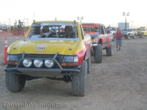 1990 Ford Bronco - yellow and red off-road race truck with a triple-light front bumper leads other trucks on a dirt lot at dusk.