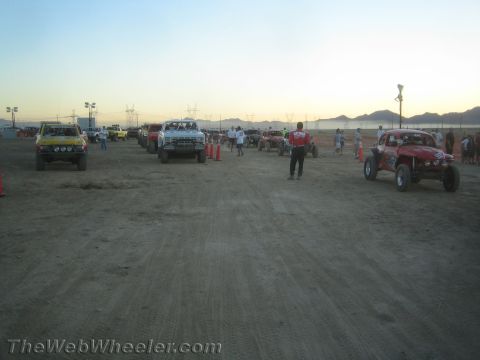 1990 Ford Bronco - Off-road race trucks and buggies staged across a wide dirt lot at sunset with desert mountains in the distance.