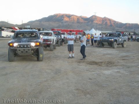1990 Ford Bronco - People stand near a line of off-road race trucks on dirt with sunlit desert mountains in the background.