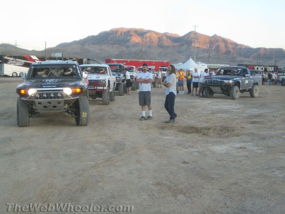People standing near off-road race trucks with sunlit mountains behind