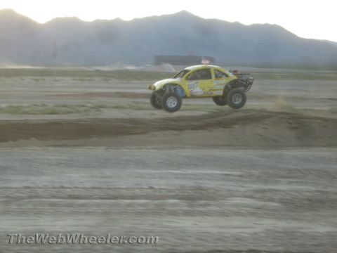 1990 Ford Bronco - yellow off-road race truck catches air over dirt mounds on a short course with desert mountains behind.